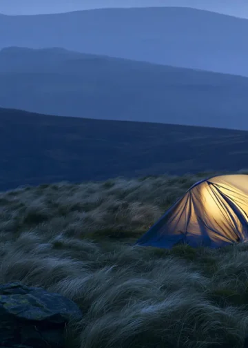 Wild camper pitching a tent on a remote UK hillside at dusk